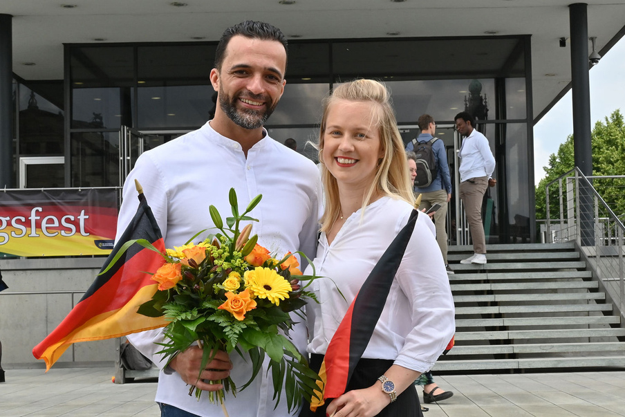 Ein Mann und eine Frau stehen mit Deutschlandfahnen un d einem Blumenstrauß vor dem Eingang zum Landtag und lächeln vor Freude.